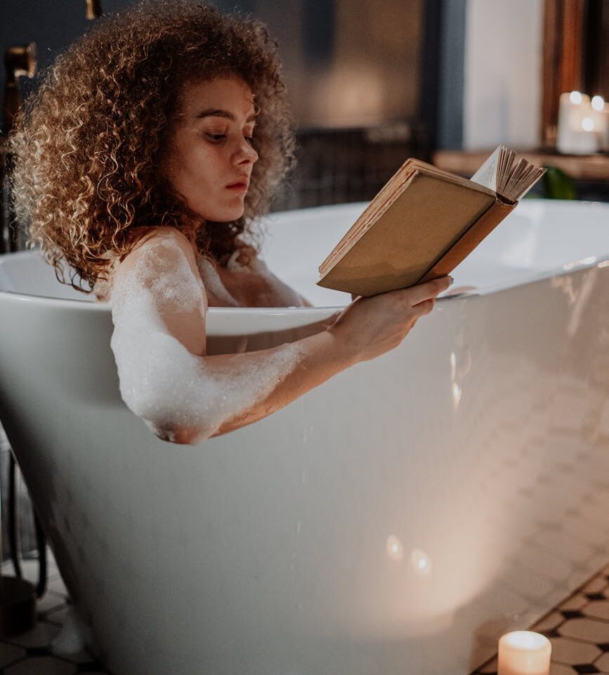 woman taking time for self care, reading a book and enjoying a bubble bath.