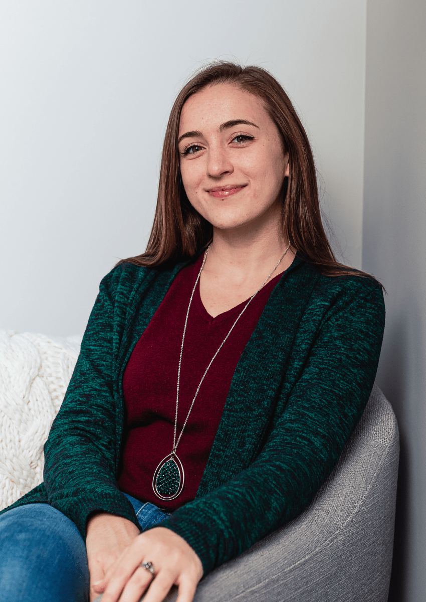 Morgan Barbret, certified Wayfinder life coach sitting in a gray armchair smiling at the camera. She is sitting upright and has one leg crossed over the other and hands together.
