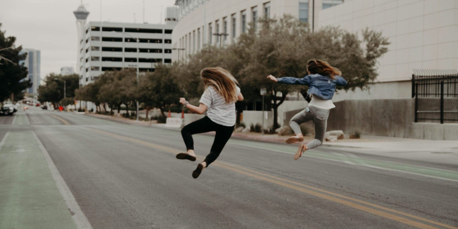 two girls walking down a paved city street, jumping into the air and clicking their heels