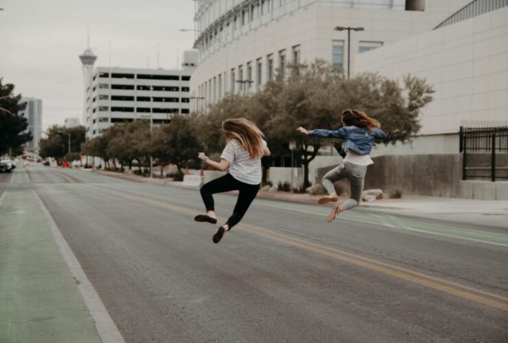 two girls walking down a paved city street, jumping into the air and clicking their heels